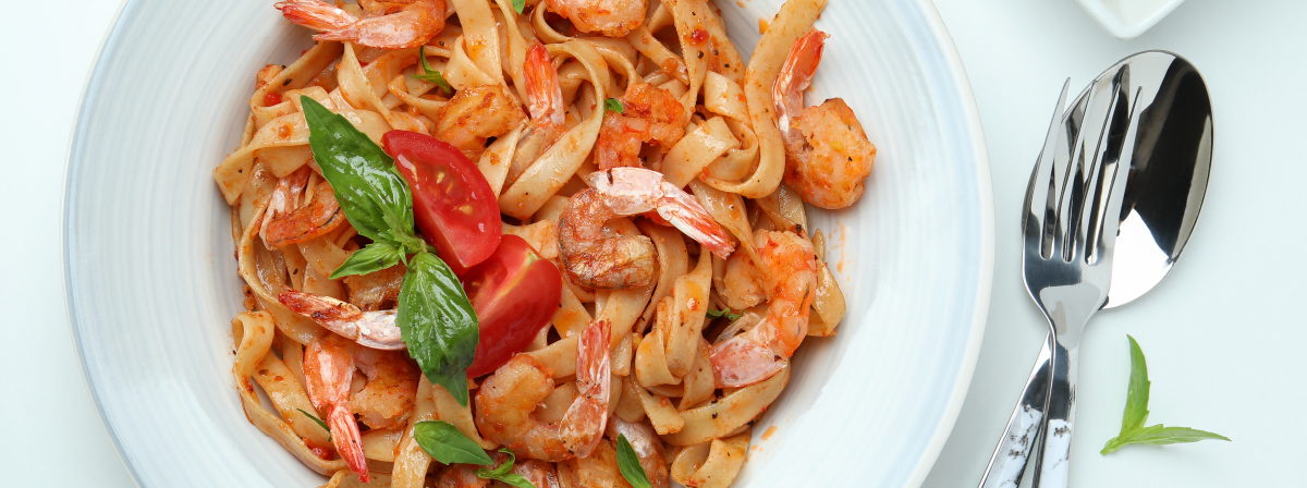 A plate of fettuccine pasta with shrimp, garnished with slices of fresh tomato and basil leaves. The dish is seasoned and appears to be tossed in a light, possibly tomato-based sauce. The plate is set on a table with a fork and a knife placed on the right side, with a small plate containing a lemon wedge in the background