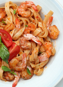 A plate of fettuccine pasta with shrimp, garnished with slices of fresh tomato and basil leaves. The dish is seasoned and appears to be tossed in a light, possibly tomato-based sauce. The plate is set on a table with a fork and a knife placed on the right side, with a small plate containing a lemon wedge in the background