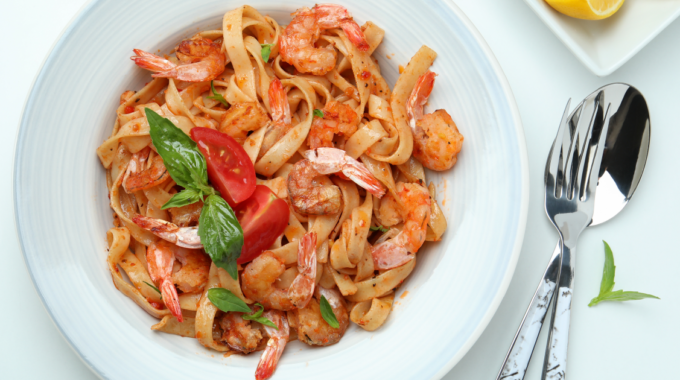 A plate of fettuccine pasta with shrimp, garnished with slices of fresh tomato and basil leaves. The dish is seasoned and appears to be tossed in a light, possibly tomato-based sauce. The plate is set on a table with a fork and a knife placed on the right side, with a small plate containing a lemon wedge in the background