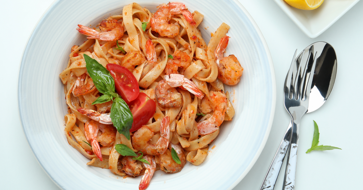 A plate of fettuccine pasta with shrimp, garnished with slices of fresh tomato and basil leaves. The dish is seasoned and appears to be tossed in a light, possibly tomato-based sauce. The plate is set on a table with a fork and a knife placed on the right side, with a small plate containing a lemon wedge in the background